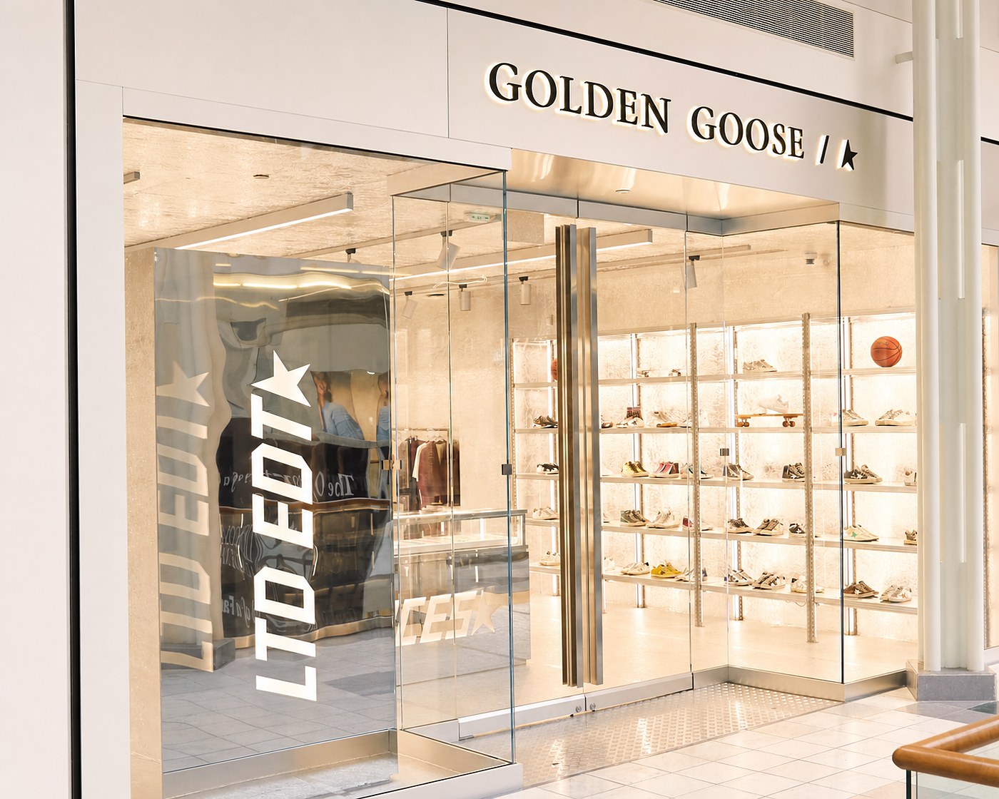 Golden Goose boutique storefront in a mall corridor, its backlit wall of sneakers glowing warm behind full-height glass